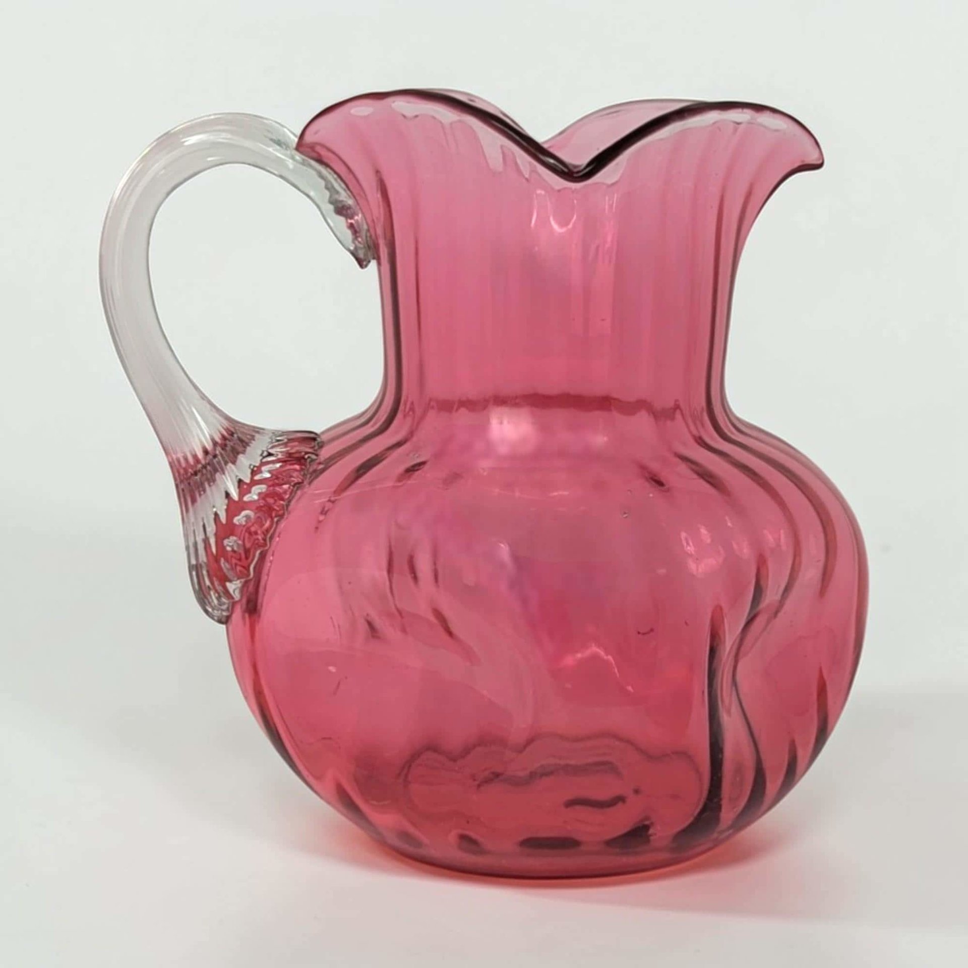 A vibrant cranberry red glass pitcher with a rounded body and a flared, ruffled rim. It features a clear glass handle attached to the side and a polished pontil mark on the bottom. The glass has a rich, translucent glow.