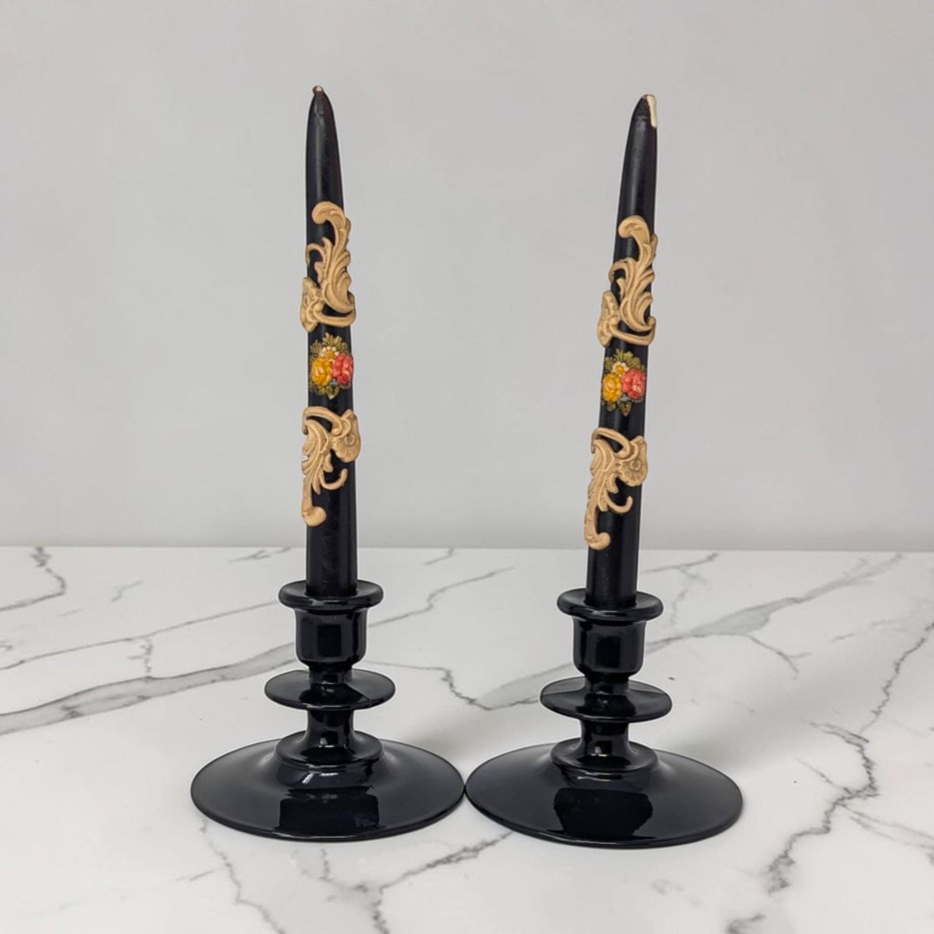 A pair of glossy black glass candle holders with a wide, tiered circular base. They hold two tall, cream-colored candles. The glass appears solid black but would show deep purple under strong light.