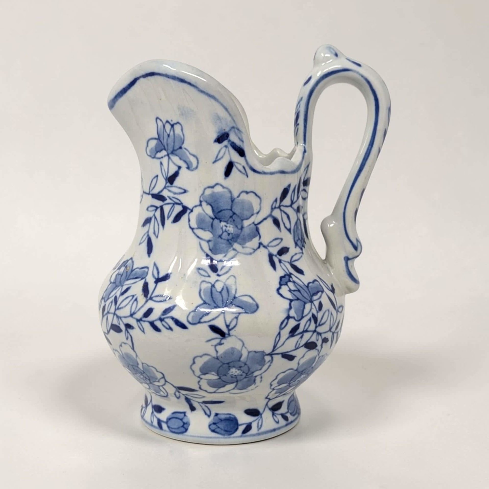 A 6-inch ceramic pitcher with a bulbous silhouette and a smooth glazed texture. It features hand-painted blue floral vine patterns on a white background with a scalloped rim and an ornate curved handle.