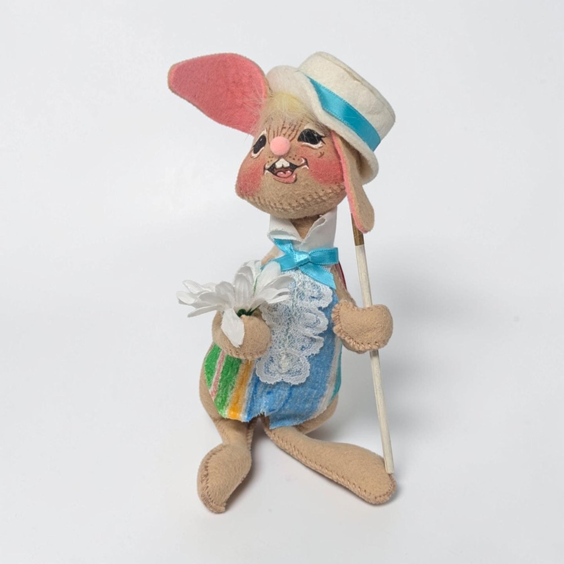 Tan felt bunny wearing a white boater hat with a blue ribbon and a striped vest in blue, yellow, and green. He holds a white daisy in one hand and a white cane in the other. He has a toothy hand-painted smile and pink-lined ears.