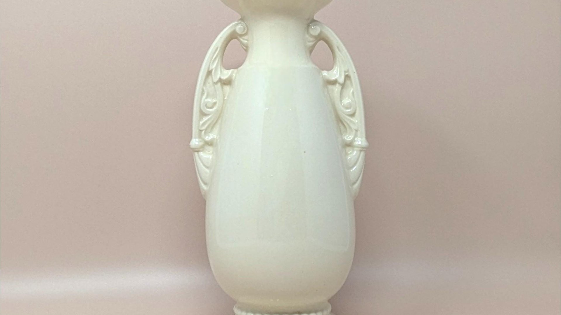 A studio photo of a tall, cream-colored ceramic vase with a glossy finish. The vase features an elegant urn shape with two ornate, scroll-style handles on the sides. It has a flared rim and sits on a tiered circular base with a beaded detail around the bottom.