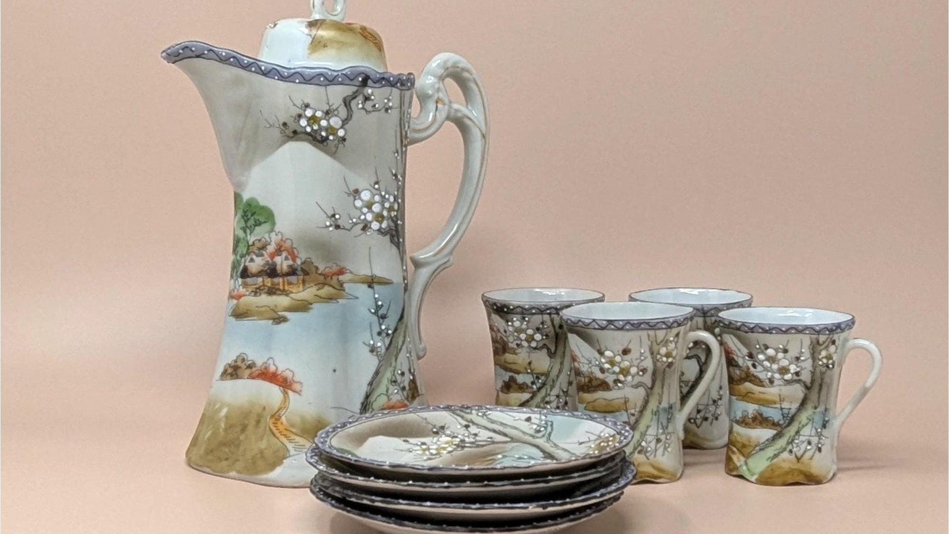 A hand-painted porcelain tea set featuring a tall teapot, four cups, and four saucers. The set depicts a traditional Japanese landscape with mountains, water, and flowering trees in soft earth tones and blue borders.