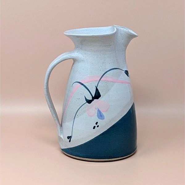 Speckled cream earthenware pitcher with a wide flared rim and sturdy handle. Features a hand-painted abstract floral design in teal, pink, and indigo with a diagonal teal color-block base.