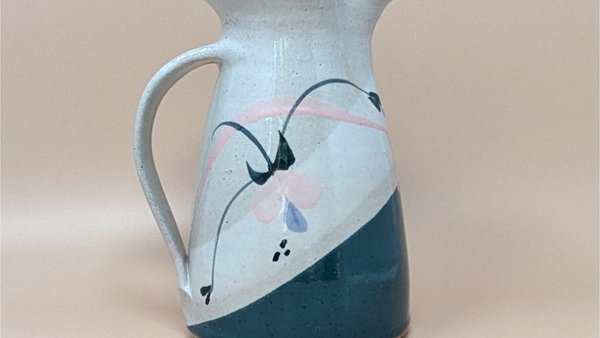 Speckled cream earthenware pitcher with a wide flared rim and sturdy handle. Features a hand-painted abstract floral design in teal, pink, and indigo with a diagonal teal color-block base.