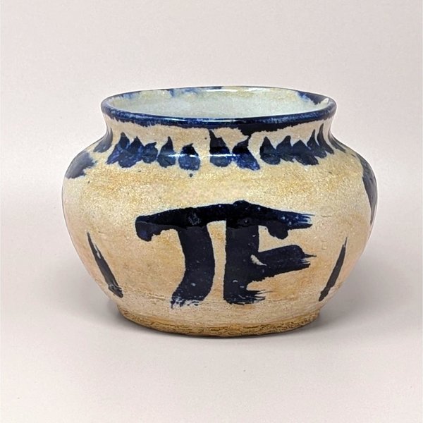 Hand-thrown pottery bowl with a globular shape and a sandy, textured tan glaze. Featuring bold, gestural cobalt blue brushstrokes including a large "PI" or "TF" symbol and a saw-tooth rim pattern.