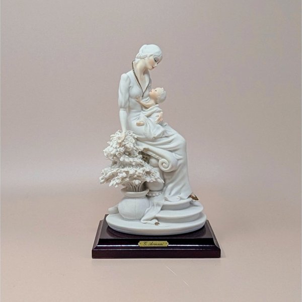 Giuseppe Armani "Maternal" porcelain figurine of a mother cradling an infant, seated beside a flowering tree. Set on a dark wood square pedestal base with a gold nameplate. Matte white and cream finish.