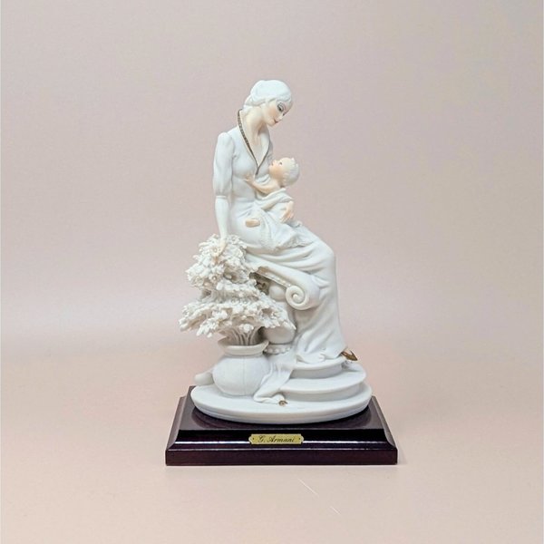 Giuseppe Armani "Maternal" porcelain figurine of a mother cradling an infant, seated beside a flowering tree. Set on a dark wood square pedestal base with a gold nameplate. Matte white and cream finish.