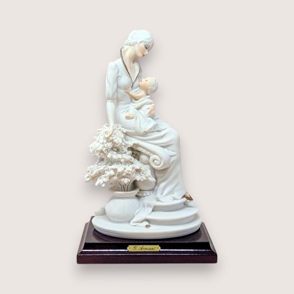 Giuseppe Armani "Maternal" porcelain figurine of a mother cradling an infant, seated beside a flowering tree. Set on a dark wood square pedestal base with a gold nameplate. Matte white and cream finish.