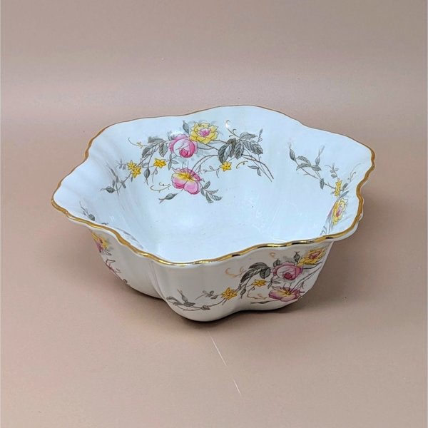 A scalloped white porcelain bowl with a gold gilt rim. Features pink and yellow rose bouquets with grey-green leaves draped across the interior and exterior. Delicate, fluted silhouette with a glossy finish on a neutral tan background.