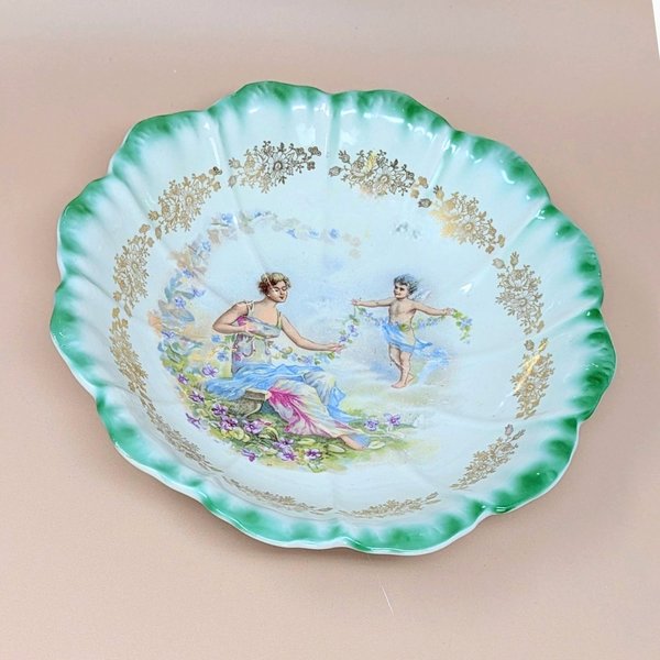 Antique Dresden porcelain bowl with a scalloped edge and green ombré rim. Center features a classical scene of a woman and a cherub with floral garlands. Gold filigree accents. 9.25 inches wide.
