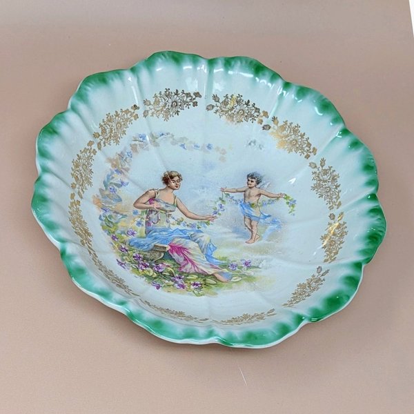 Antique Dresden porcelain bowl with a scalloped edge and green ombré rim. Center features a classical scene of a woman and a cherub with floral garlands. Gold filigree accents. 9.25 inches wide.