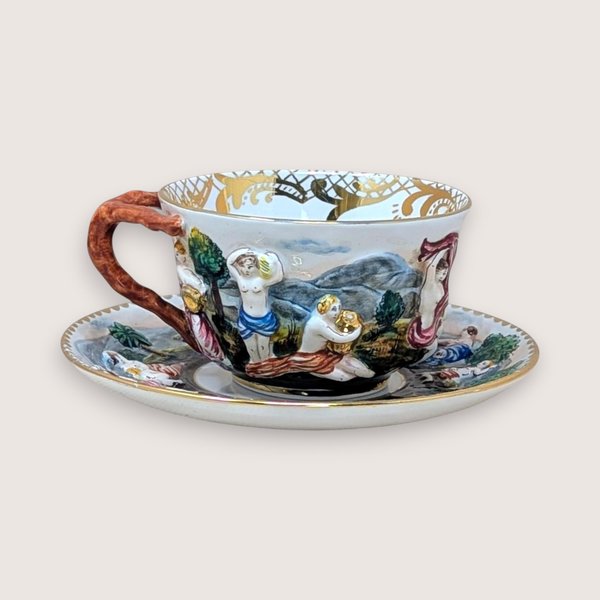Italian Capodimonte porcelain cup and saucer set. Features high-relief, hand-painted classical figures in a woodland landscape. Intricate gold gilt interior accents and a rustic coral-style handle. Cup 2.5", Saucer 6".