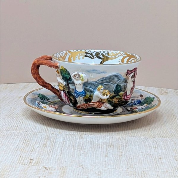 Italian Capodimonte porcelain cup and saucer set. Features high-relief, hand-painted classical figures in a woodland landscape. Intricate gold gilt interior accents and a rustic coral-style handle. Cup 2.5", Saucer 6".