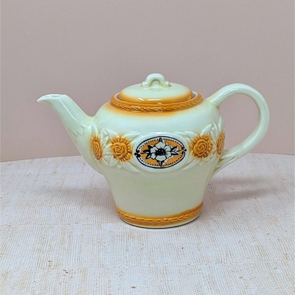 A vintage Porcelier cream-colored teapot with a raised, textured sunflower and rope border in amber-brown tones. A central black and white floral medallion sits within an oval frame. The teapot has a classic rounded shape and matching lid, set against a neutral, warm background.