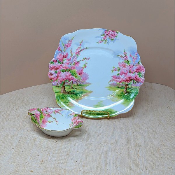 Two-piece Royal Albert Blossom Time bone china set. Includes a 9.75-inch square handled cake plate and a 5.5-inch oval handled sweet meat dish. Features pink blossom trees and gold-trimmed scalloped edges.