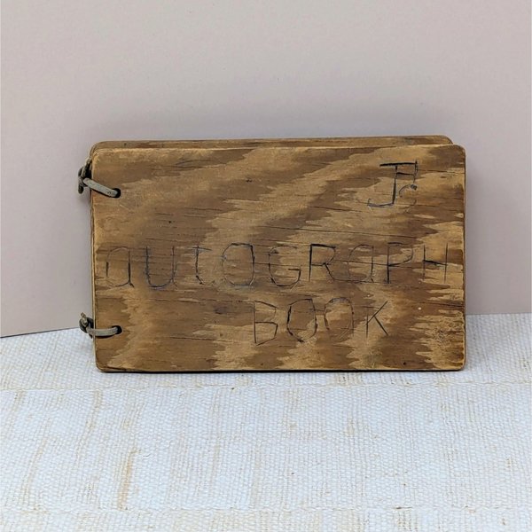 A small, rectangular autograph book with a natural wood grain cover. The words "Autograph Book" are hand-carved into the wood. Two metal rings bind the pages on the left. Rustic, weathered texture with a warm honey-oak finish.