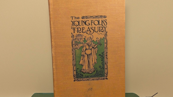 An antique hardcover book with a gold-toned textured cloth binding. A central green and black illustration shows a medieval-style woman with a falcon on her hand, framed by intricate Art Nouveau borders.