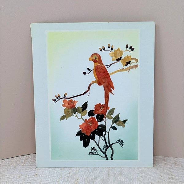 A vertical rectangular wall art piece featuring a bird perched on a branch, meticulously crafted from layered bamboo. Warm sienna and ochre tones are accented by deep red floral blooms. Framed with a clean white and pink double mat.