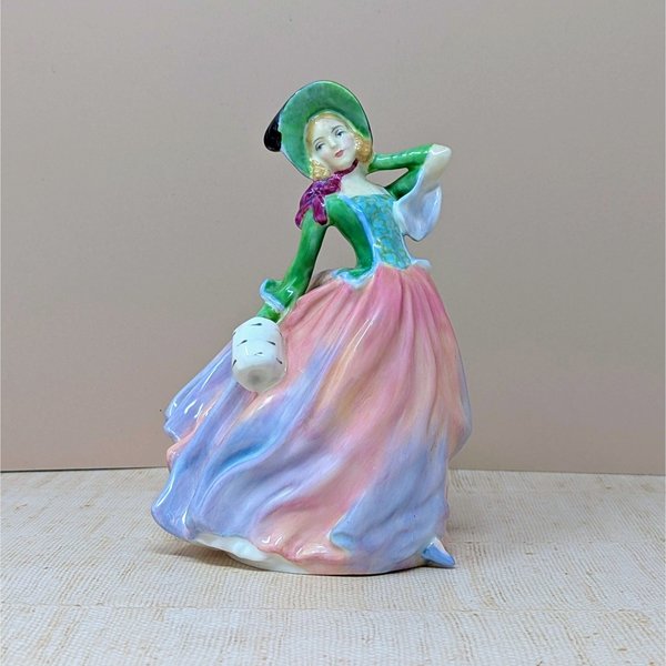 A vertical porcelain figurine of a woman in a wind-swept gown. She wears a vibrant green bodice, a wide-brimmed hat, and a voluminous skirt in blended shades of pink, peach, and lavender. Glossy, fine-bone china texture.