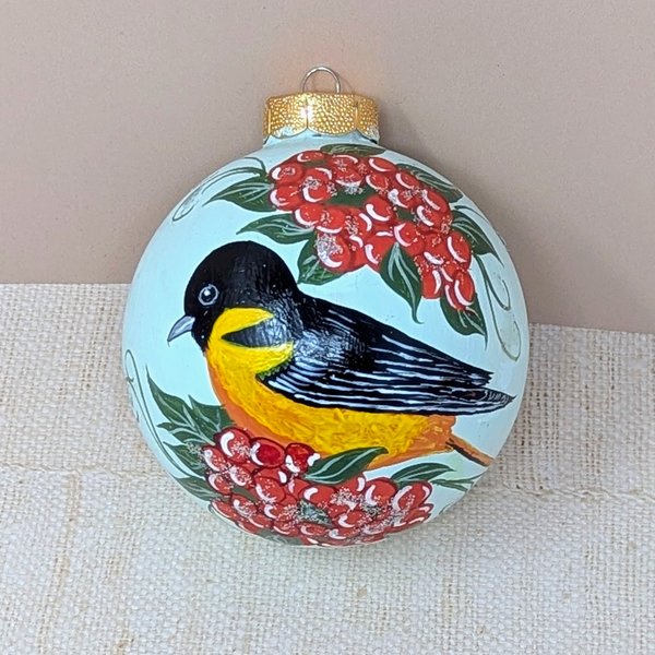 A spherical glass ornament featuring a hand-painted Baltimore Oriole bird with a black head and vibrant orange breast. Surrounded by clusters of bright red berries and green leaves. Topped with a classic gold metal cap.