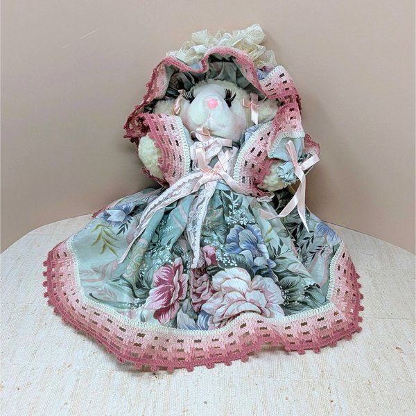A seated plush lamb in a wide, floral-patterned sage green and rose pink dress. The lamb features long eyelashes, a pink nose, and an oversized matching lace bonnet. The intricate pink lace trim on the hem and bonnet adds a grounded, romantic texture.