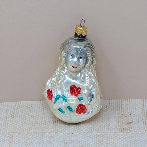 A vintage mercury glass ornament featuring a Victorian-style girl with long, flowing hair holding a bouquet of red roses with green leaves. The glass has a warm, milky-white luster with hand-painted facial details.