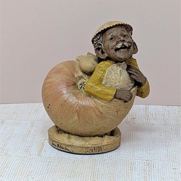 A hand-cast resin sculpture of a smiling gnome-like character named Sandy emerging from a large peach-colored seashell. The character wears a yellow jacket and a small hat. 4.75 inches tall.