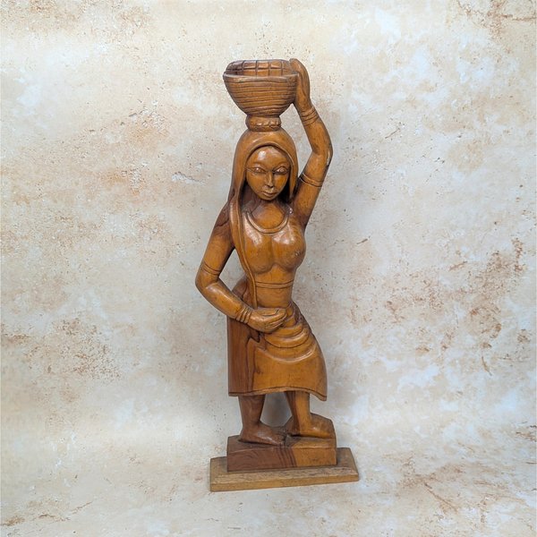 A vintage mid-century hand carved wooden statue of an African woman on a tan textured background. Standing twenty four inches tall, the solid timber figure gracefully balances a woven basket on her head with one arm raised.