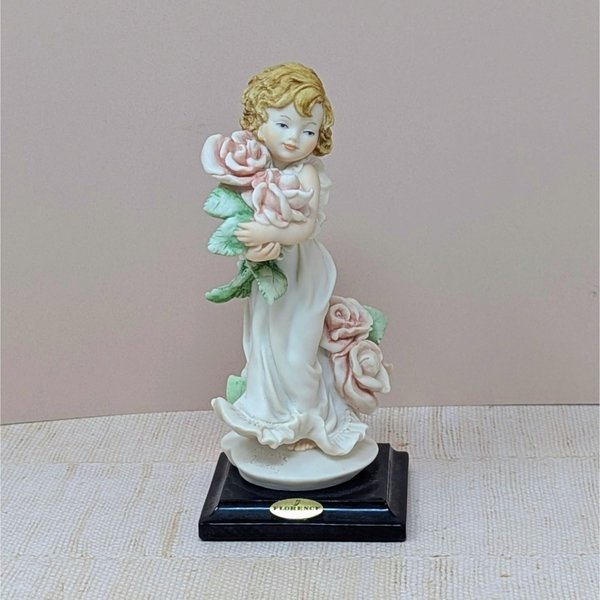 A finely detailed porcelain figurine of a young girl in a flowing white dress holding a large bouquet of pink roses. She stands on a dark wooden base with a gold Florence Giuseppe Armani plaque.