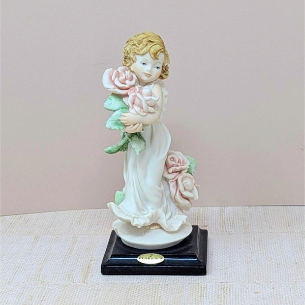 A finely detailed porcelain figurine of a young girl in a flowing white dress holding a large bouquet of pink roses. She stands on a dark wooden base with a gold Florence Giuseppe Armani plaque.