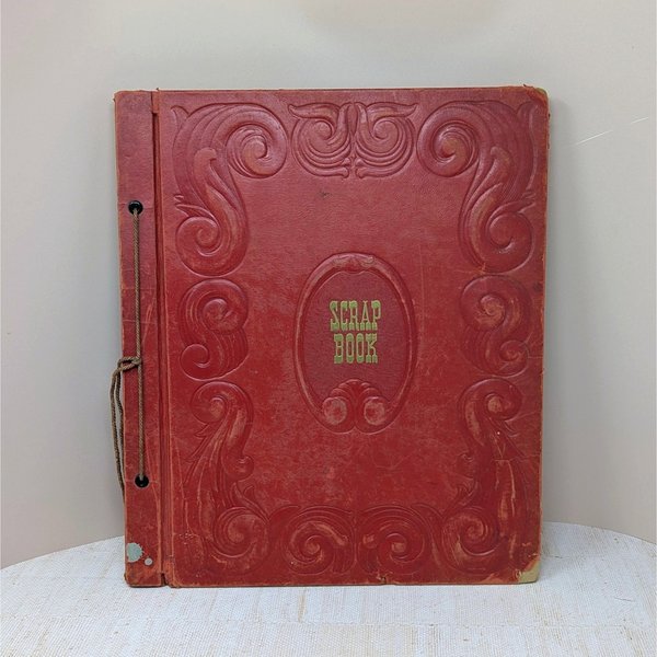 A large, vintage red leatherette scrapbook with an ornate embossed border and "Scrapbook" in gold lettering at the center. The cover shows significant age-related wear and is secured by a brown cord binding.