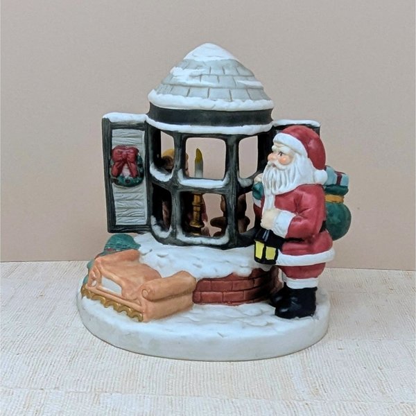 A painted ceramic music box featuring santa standing by a snowy bay window. A small child is visible inside the house through the panes. The colors are classic red, evergreen, and snowy white with a matte finish.