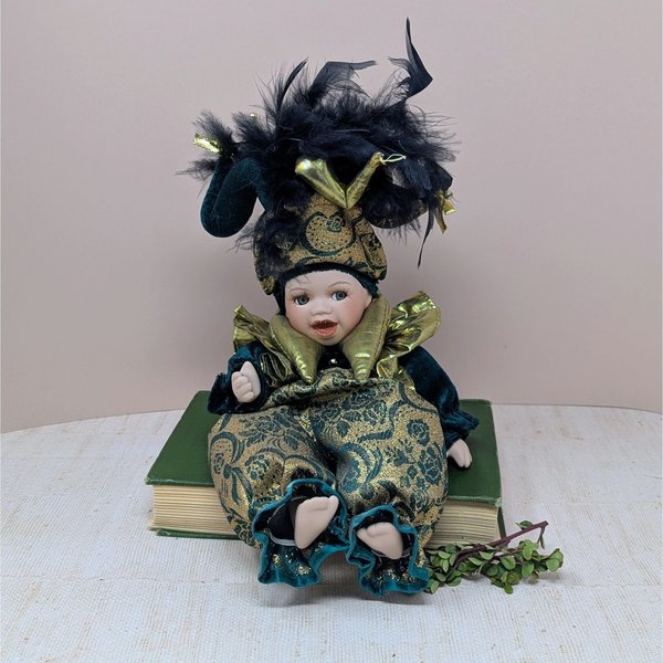 A 12-inch porcelain baby doll dressed as a jester in emerald green velvet and gold brocade, featuring a large feathered jester hat and gold metallic ruff.