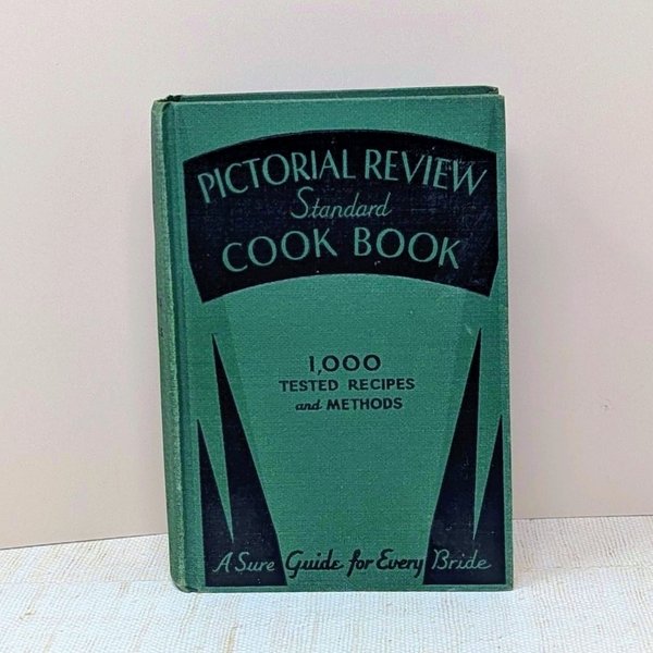 A small hardcover book with a sage green cloth cover. It features a bold black art deco design with sharp geometric points. The title Pictorial Review Standard Cook Book is printed in a stylized vintage font.