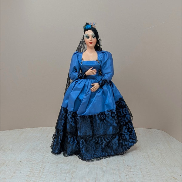 A vintage 13-inch Spanish souvenir doll featuring a cobalt blue satin dress with black lace tiers, a matching black lace mantilla veil, and dramatic hand-painted facial features against a neutral background.