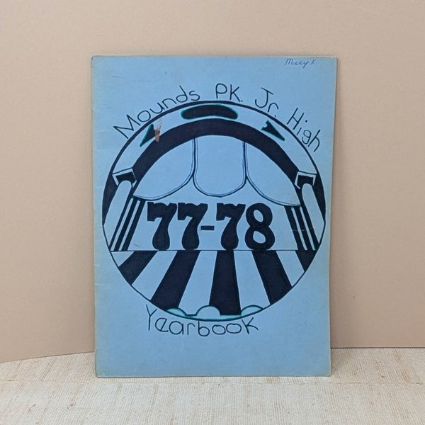 A vintage softcover yearbook featuring a light blue cover with a hand-drawn circular graphic in black ink. The center shows the bold numbers 77-78 under a theatrical or arched motif. A name is written in small ink at the top right.