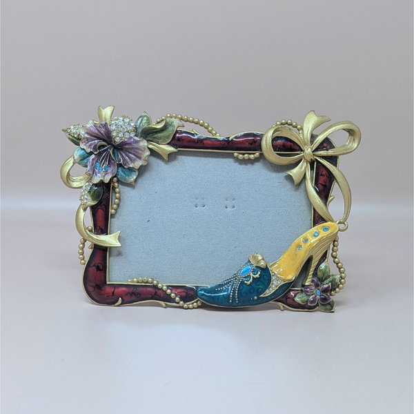 A horizontal photo frame with a deep red enamel border. Ornate gold-tone decorations include a crystal-studded purple hibiscus-style flower on the top left and a fashionable stiletto shoe on the bottom right.