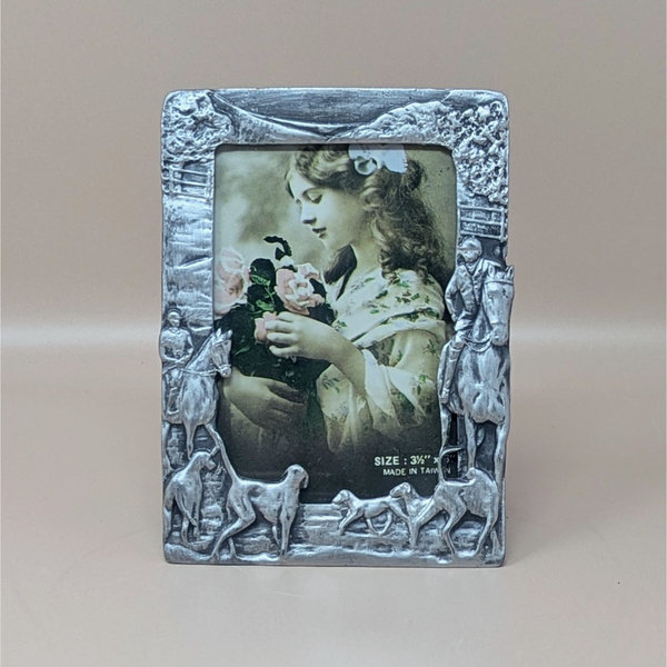 A vertical cast-metal picture frame with a weathered silver finish. Embossed hunters on horses and a pack of running hounds frame a portrait of a young girl with flowers.