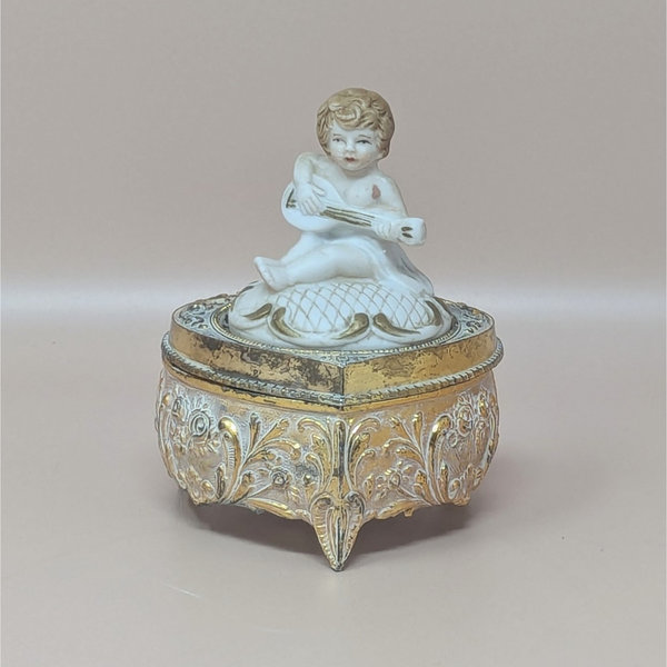 A vintage footed trinket box with a highly detailed gold-toned floral base and a white bisque-style lid featuring a cherub playing a stringed instrument.