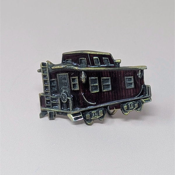 A vintage 1976 belt buckle shaped like a train caboose with deep red enamel and brass-toned metal accents on a neutral background.