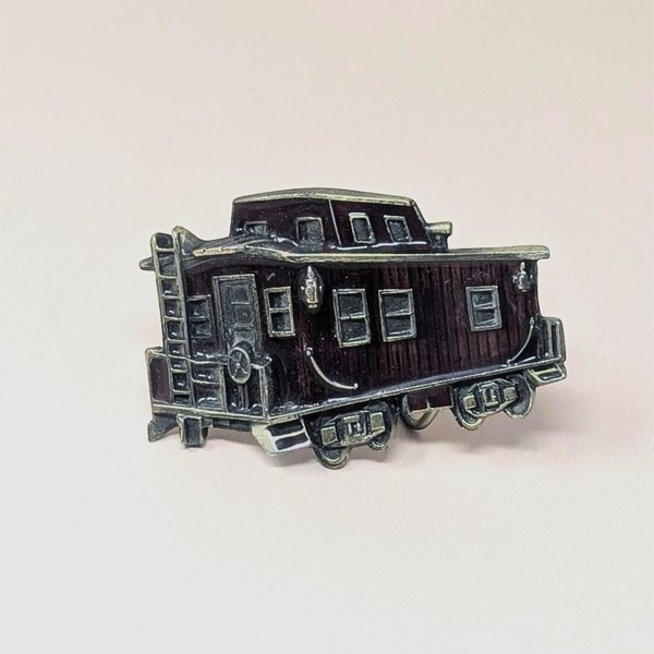 A vintage 1976 belt buckle shaped like a train caboose with deep red enamel and brass-toned metal accents on a neutral background.