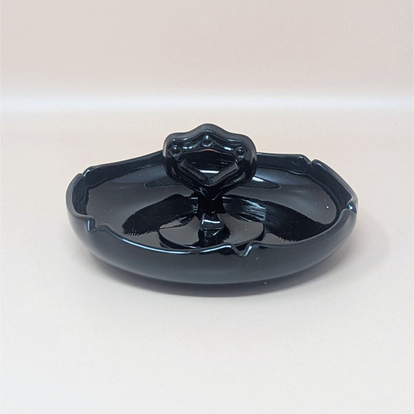 Glossy black amethyst glass trinket dish with a scalloped perimeter and a unique, ornate center handle featuring circular cutouts. The deep violet undertones are visible only under intense light.