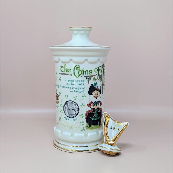 A tall white porcelain decanter featuring colorful Irish coin graphics, a leprechaun illustration, and a gold-trimmed harp stopper against a neutral background.
