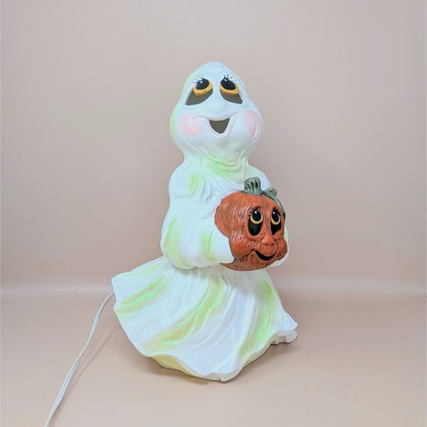 A large white ceramic ghost figurine with iridescent green highlights holding an orange jack o' lantern, featuring large expressive eyes and a wide smile.