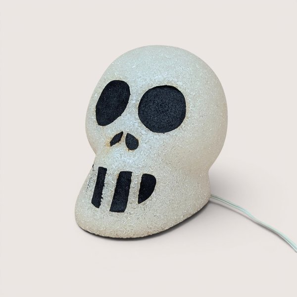 A vintage white melted plastic popcorn skull light with black painted eyes and nose sitting on a plain light background