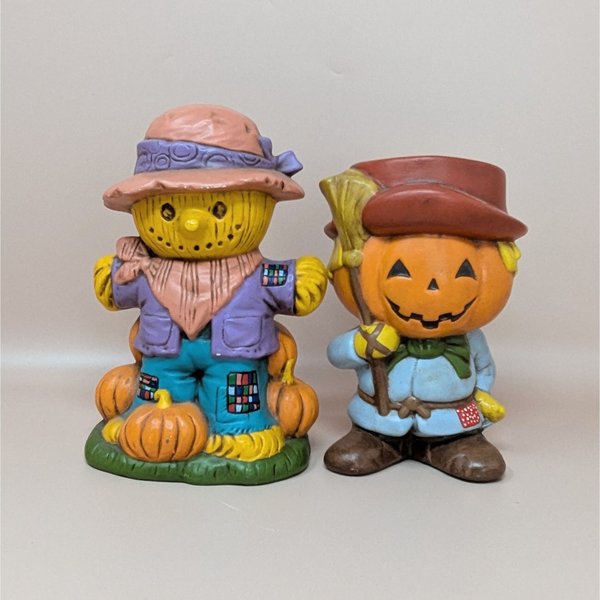 A pair of vintage hand-painted ceramic figurines featuring a friendly scarecrow standing on pumpkins and a jack o' lantern man holding a broom.