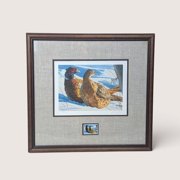 A 1987 Minnesota Pheasant Habitat Stamp Print by Brian Jarvi on a tan textured background. The framed art features a signed and numbered print of a pheasant pair in the snow, a matching stamp below, an oatmeal linen mat, and a dark wood frame.