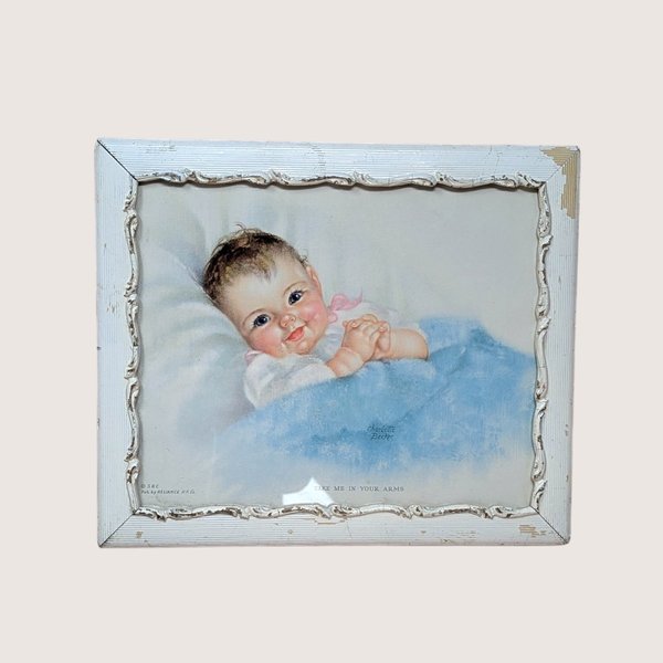 A 1940s vintage Charlotte Becker framed nursery print on a soft pink background. The sweet lithograph depicts a rosy-cheeked baby tucked under a blue blanket, housed in a distressed white decorative wooden frame with chipped paint accents.