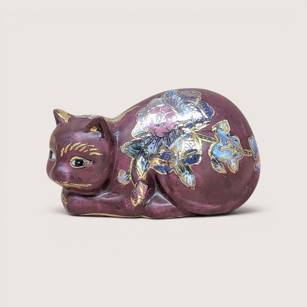 A mauve ceramic reclining cat figurine with gold and floral details, centered on a beige background.