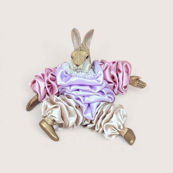 A rabbit doll in a purple and pink satin clown costume, sitting centered on a beige background.